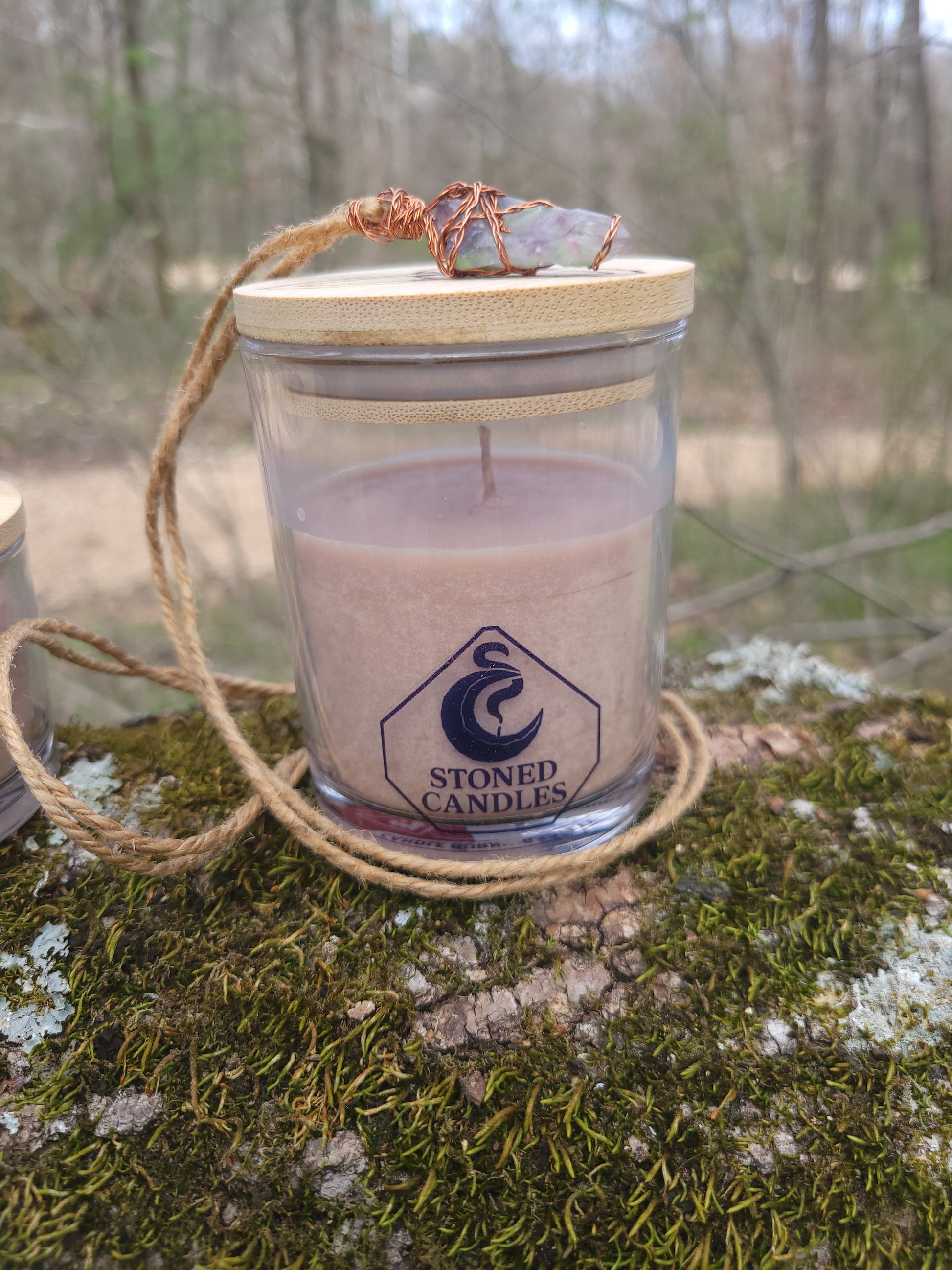 Candle in a glass jar with a wooden lid, hanging from a rope against a natural background.