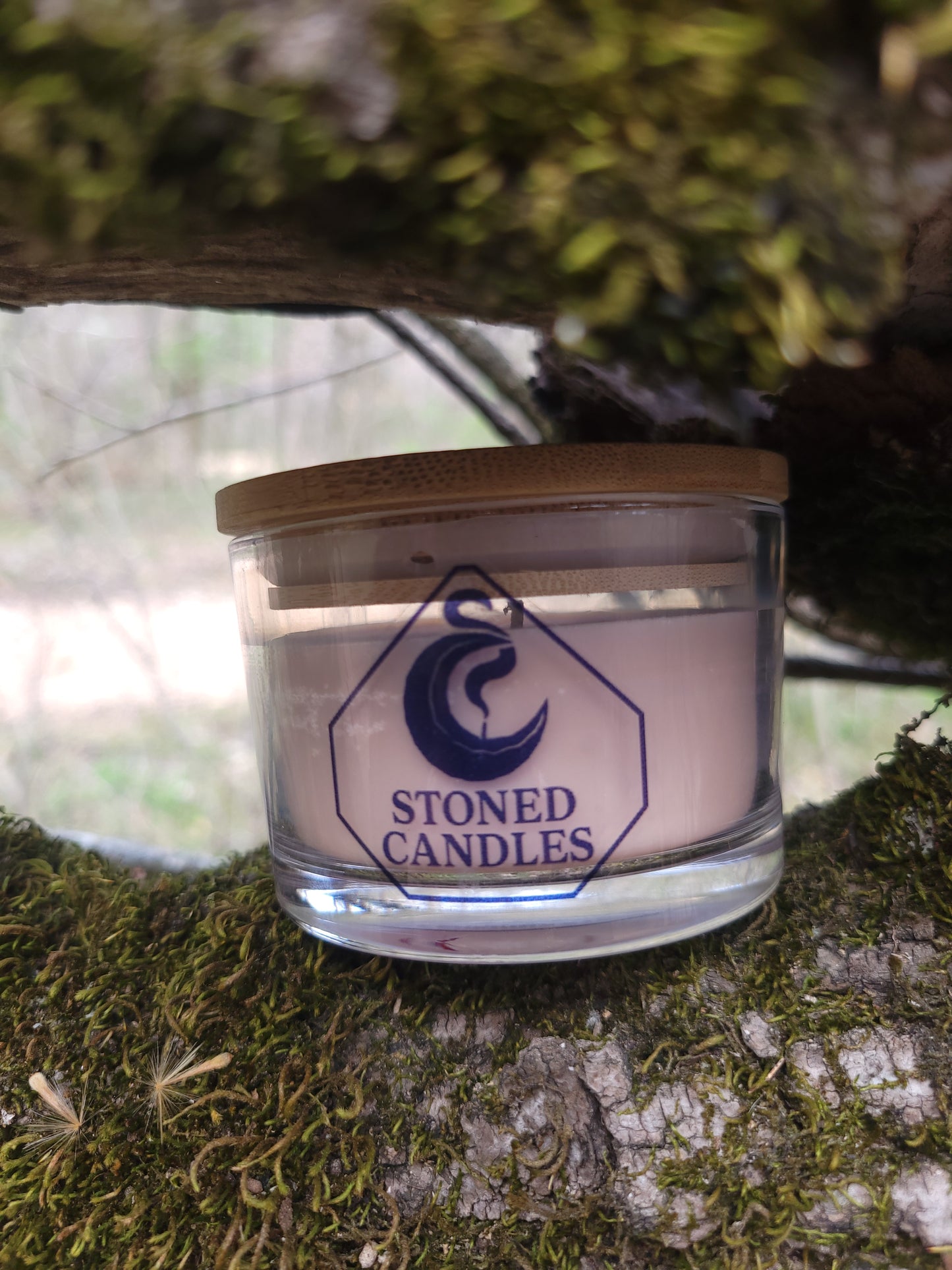 Glass candle with wooden lid on a tree trunk, featuring 'Stoned Candles' branding.