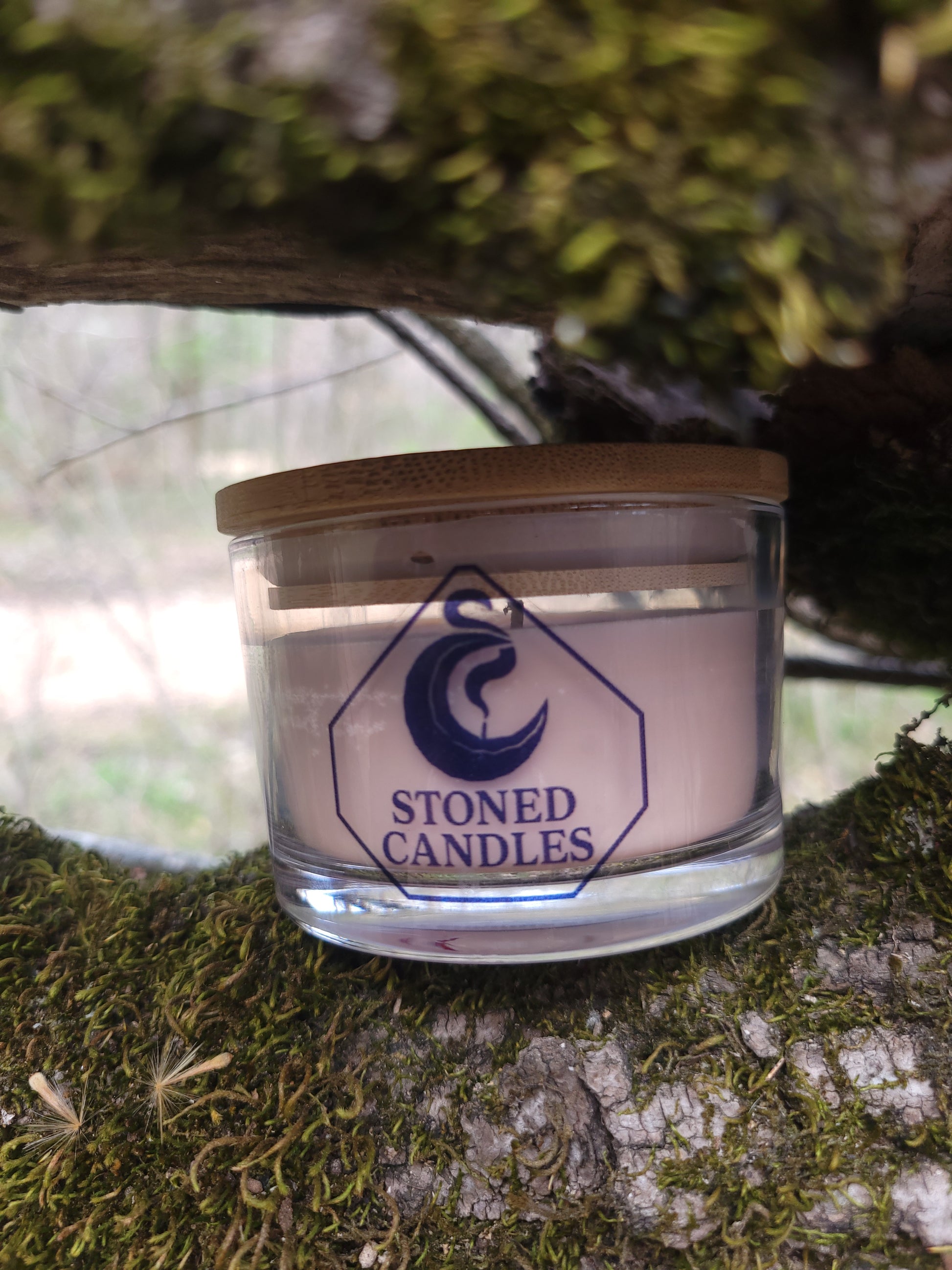 Glass candle with wooden lid on a tree trunk, featuring 'Stoned Candles' branding.