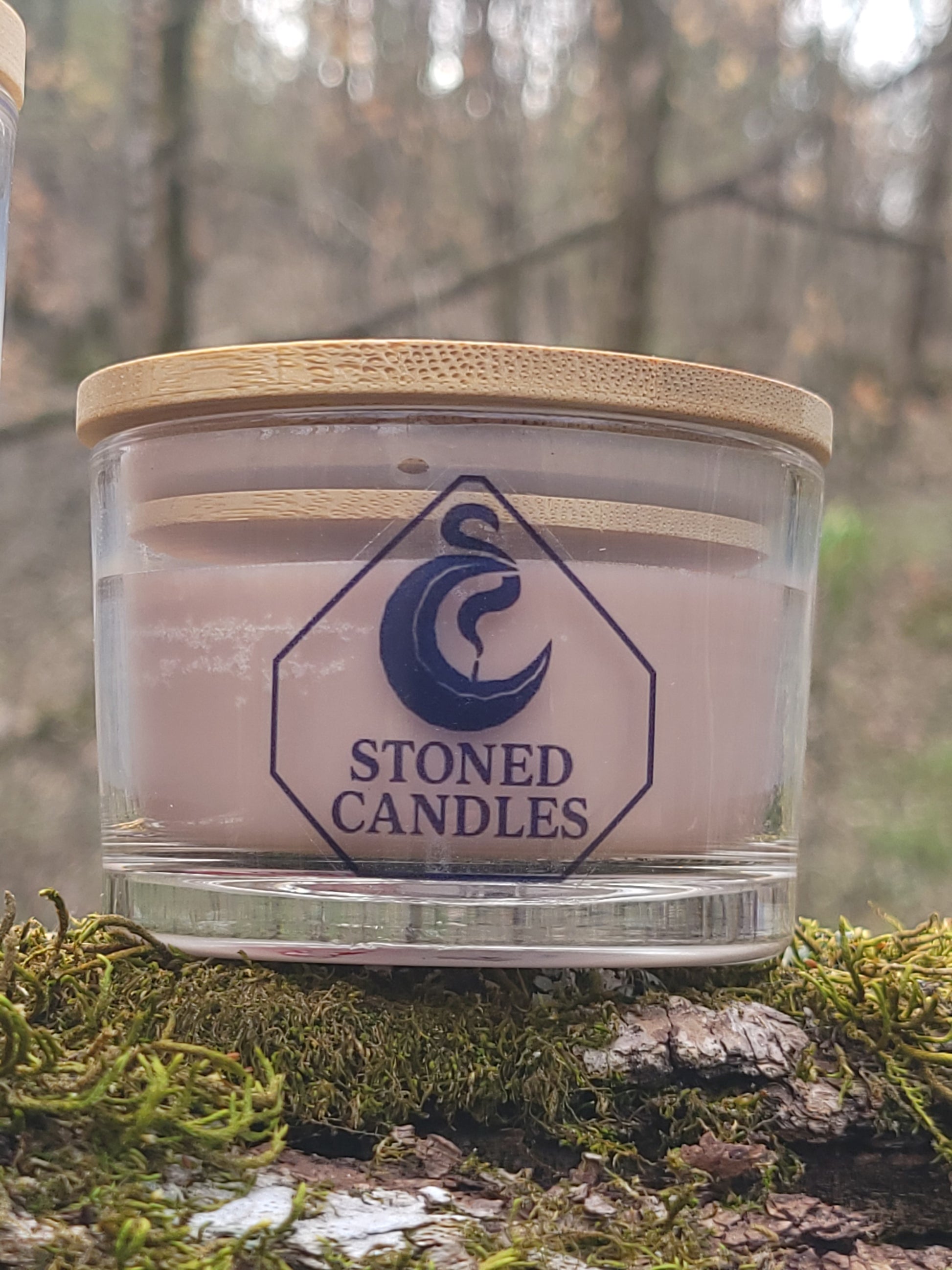 Glass candle with wooden lid on a tree branch with moss, featuring 'Stoned Candles' branding.