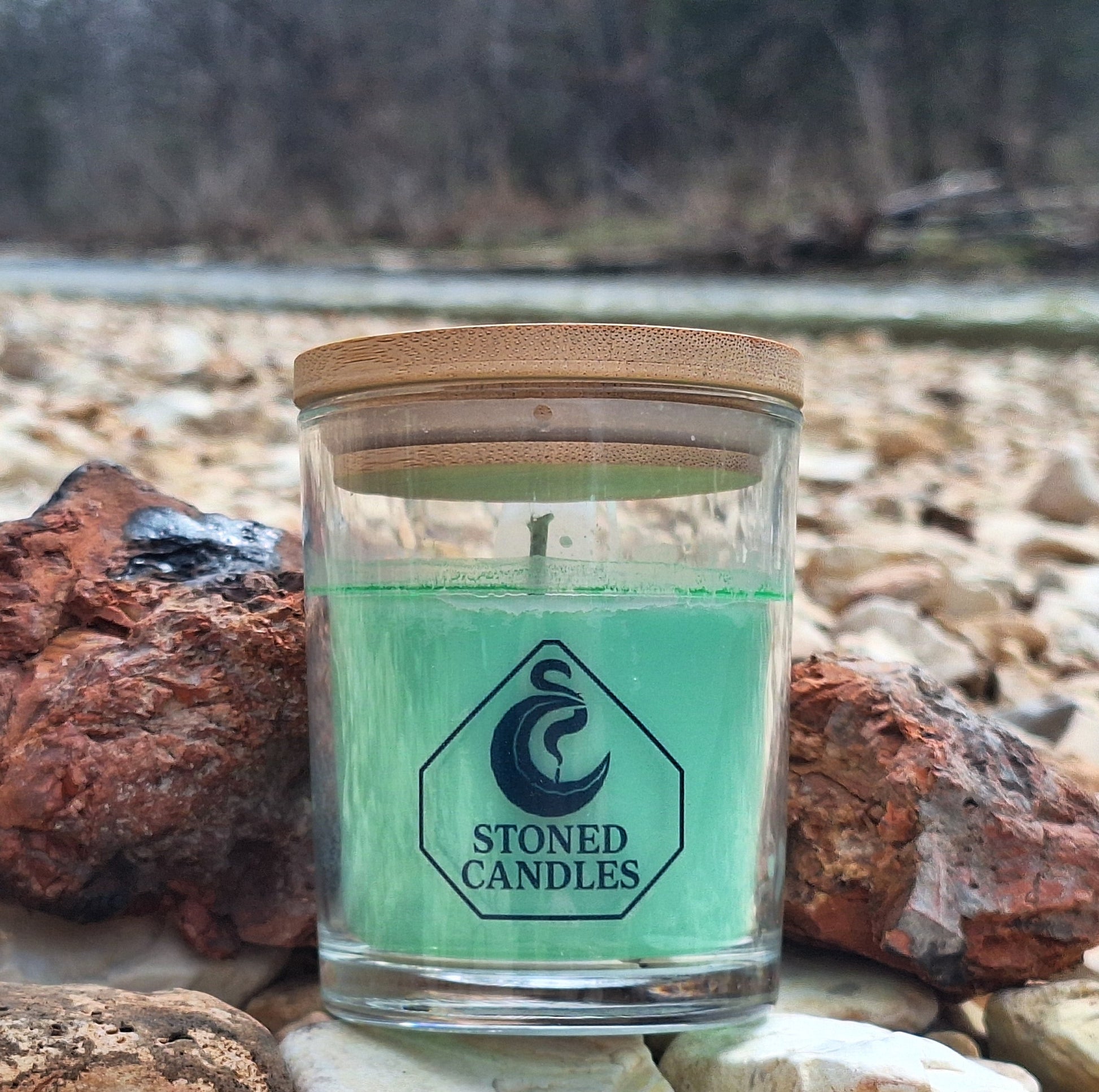 Candle in a glass jar with wooden lid on rocks near a body of water, featuring 'Stoned Candles' branding.