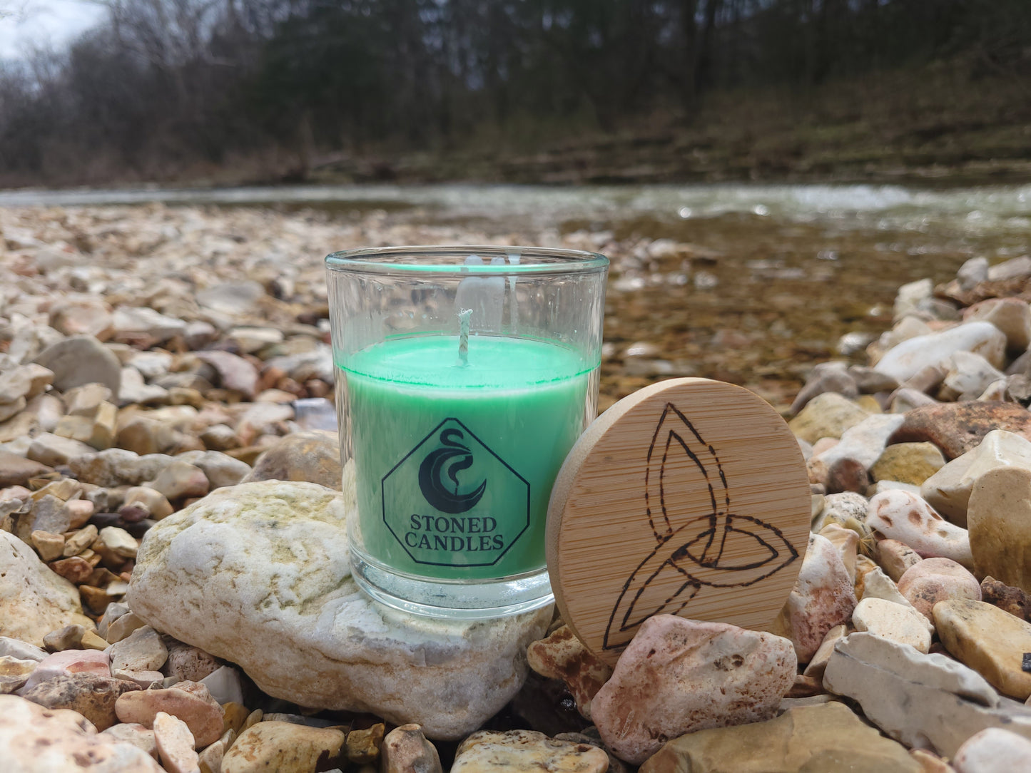 Glass candle with green wax on a stone surface, surrounded by pebbles.