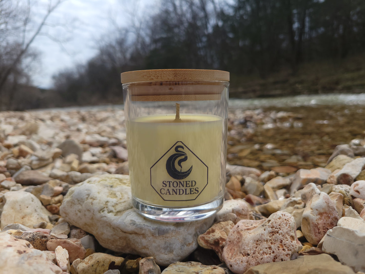 Candle in a glass jar with a wooden base, featuring a logo, on a stone surface.