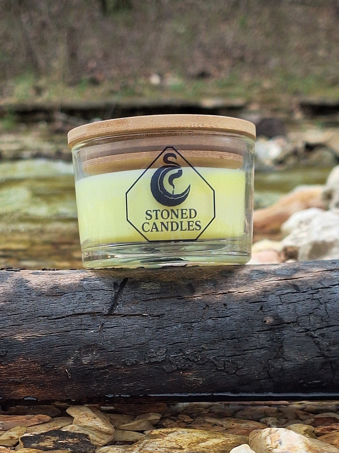 Candle jar with wooden lid labeled 'Stoned Candles' on a log by a stream.