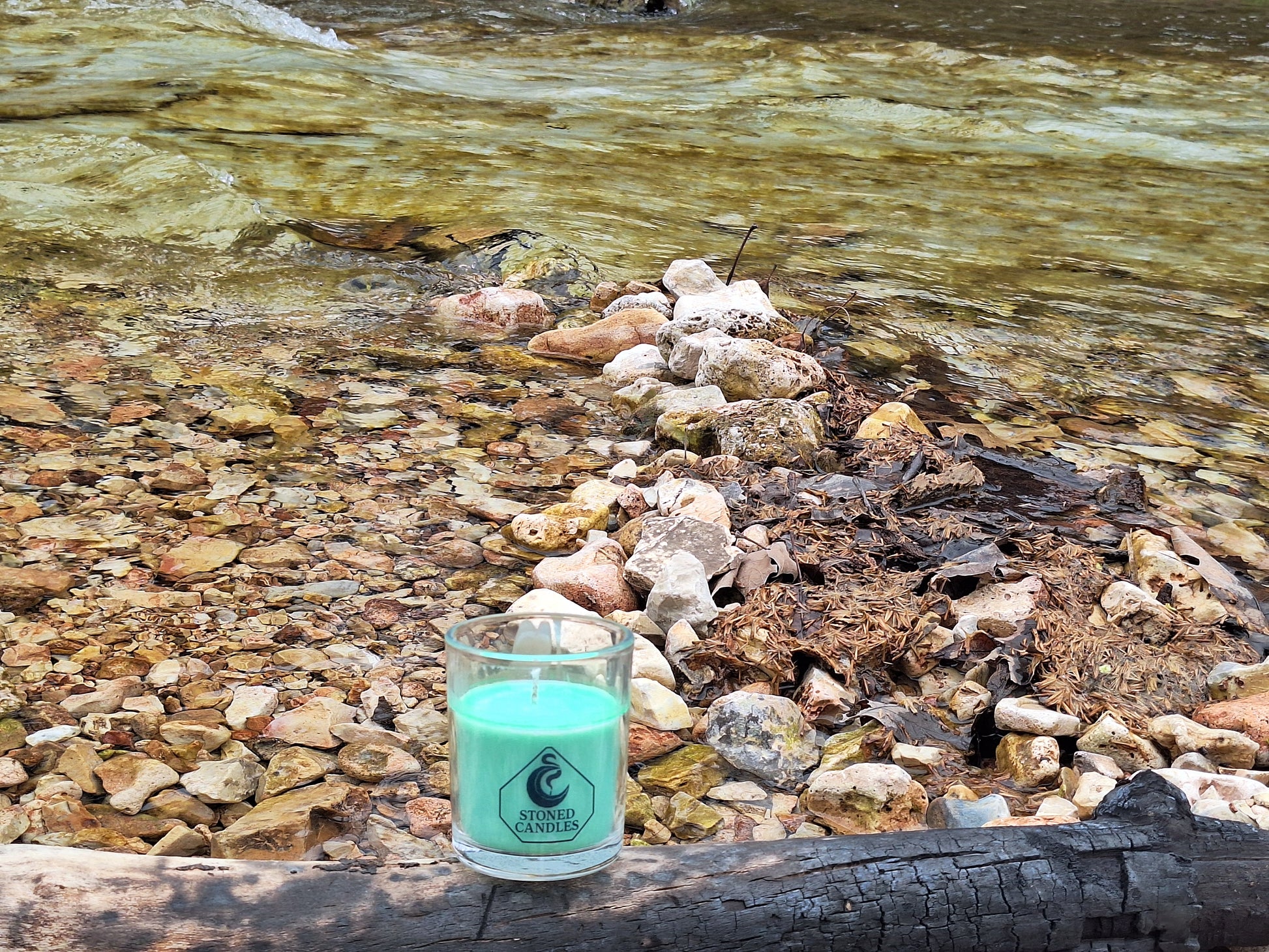 Candle in a glass jar with a visible brand logo on a rocky shoreline with water in the background
